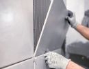Person setzt große graue Wandfliese mit Kleber ein, trägt Handschuhe, präzise Arbeit bei Fliesenverlegung sichtbar