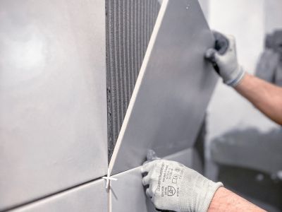 Person setzt große graue Wandfliese mit Kleber ein, trägt Handschuhe, präzise Arbeit bei Fliesenverlegung sichtbar Person setzt große graue Wandfliese mit Kleber ein, trägt Handschuhe, präzise Arbeit bei Fliesenverlegung sichtbar