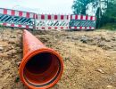 Orange Rohrleitung liegt auf sandigem Boden vor Baustellenabsperrung mit rot-weißen Barrieren.