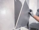 Person setzt große graue Wandfliese mit Kleber ein, trägt Handschuhe, präzise Arbeit bei Fliesenverlegung sichtbar.